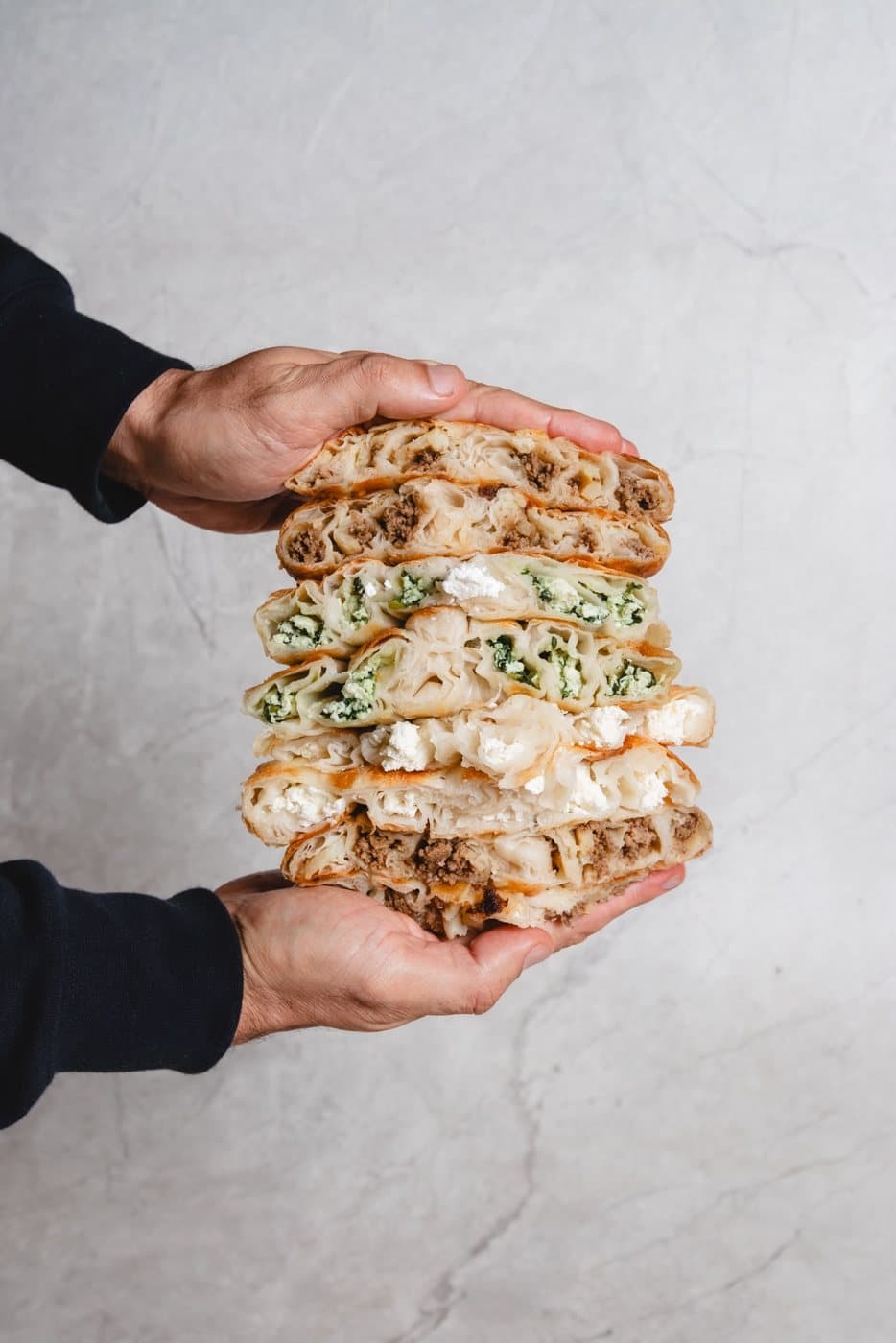 Two hands holding a stack of sliced burek, meat and spinach and cheese
